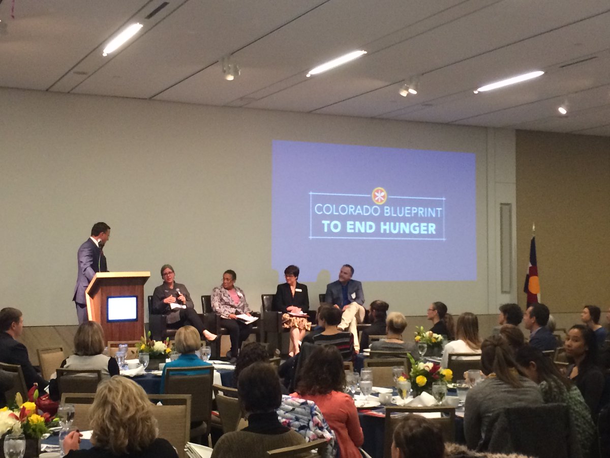 Panel of 4 (and one moderator) speak to a crowd seated at round tables. The Colorado Blueprint to End Hunger logo is projected on the wall behind them