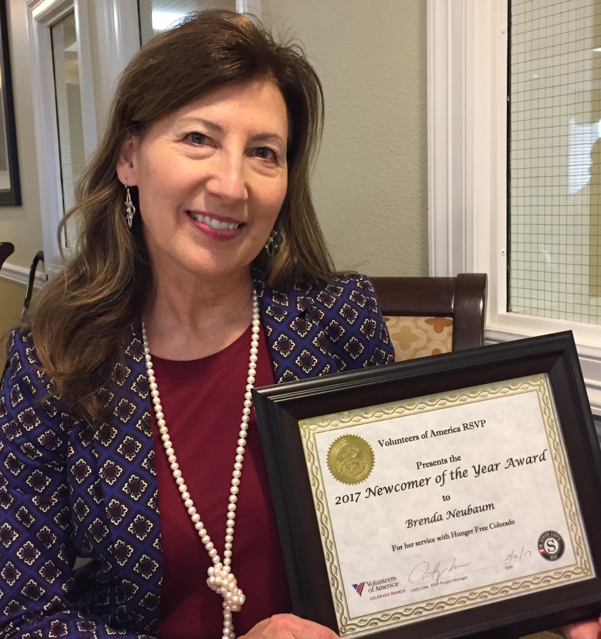 Brenda Neubaum in patterned blue blazer, long white pearl necklace and marron top, holds a black framed certificate for the 2017 Newcomer of he Year Award. Recognition is for her service to Hunger Free Colorado from the Volunteers of America RSVP.