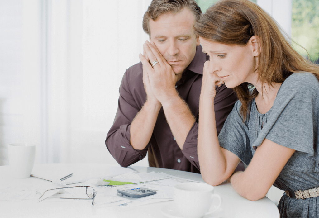 A man and a woman sit together at a table with bills, a calculator, glasses and a mug looking sad and stressed